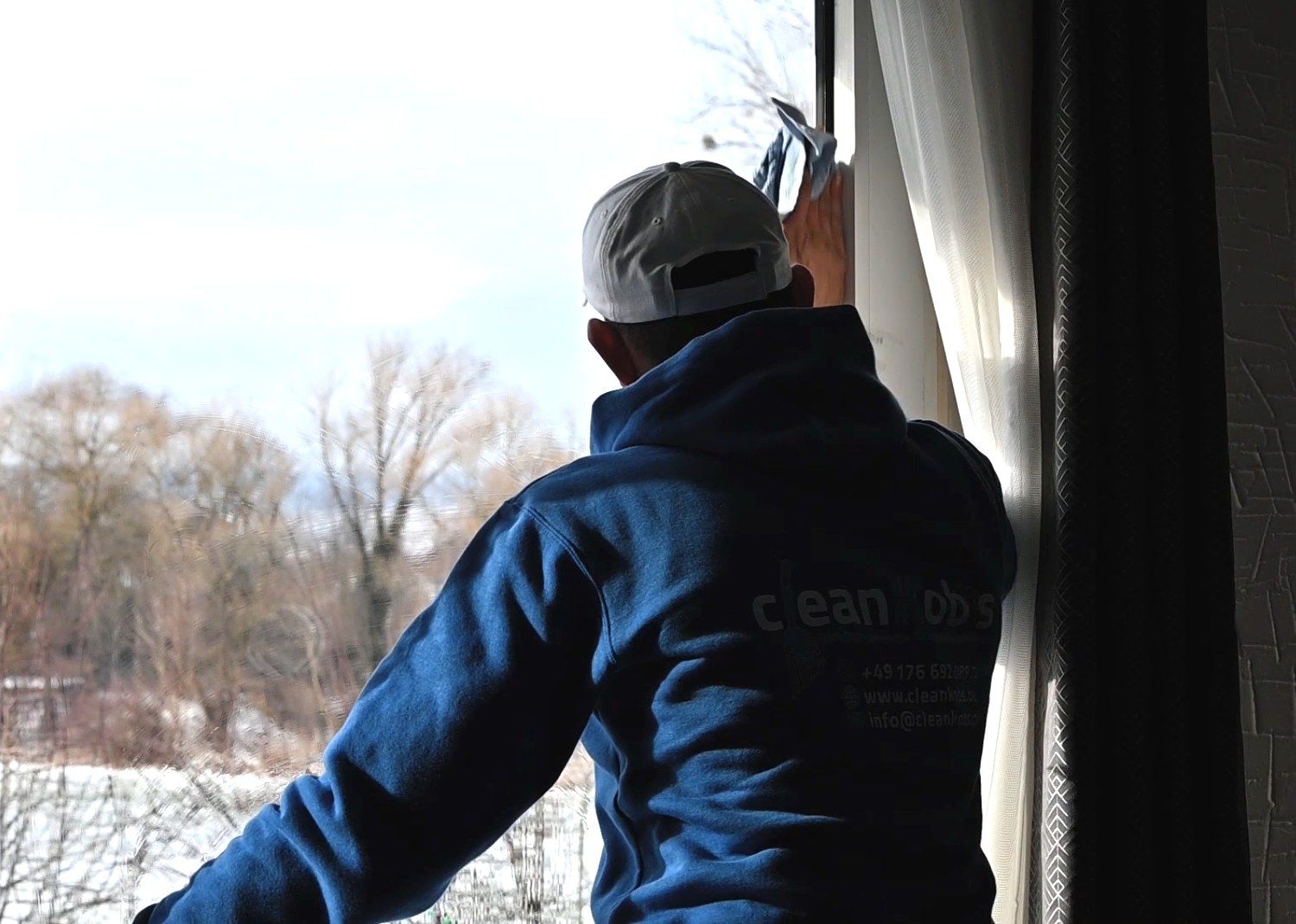 Mitarbeiter von CleanKobs im blauen Pullover mit Firmenlogo putzt ein großes Fenster mit Blick auf eine winterliche Landschaft.